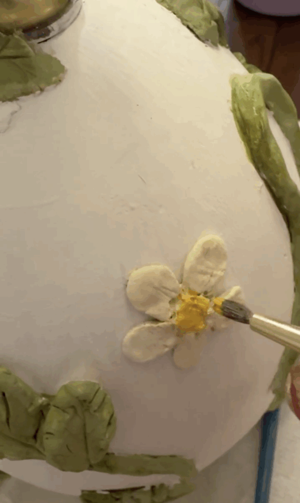 Close-up of painting a clay strawberry flower yellow on a lamp base with vines and leaves.