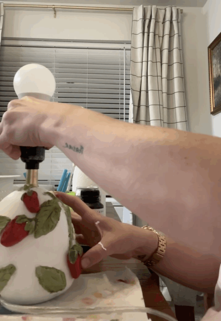 Person attaching red painted clay strawberries to a lamp decorated with green leaves and vines.