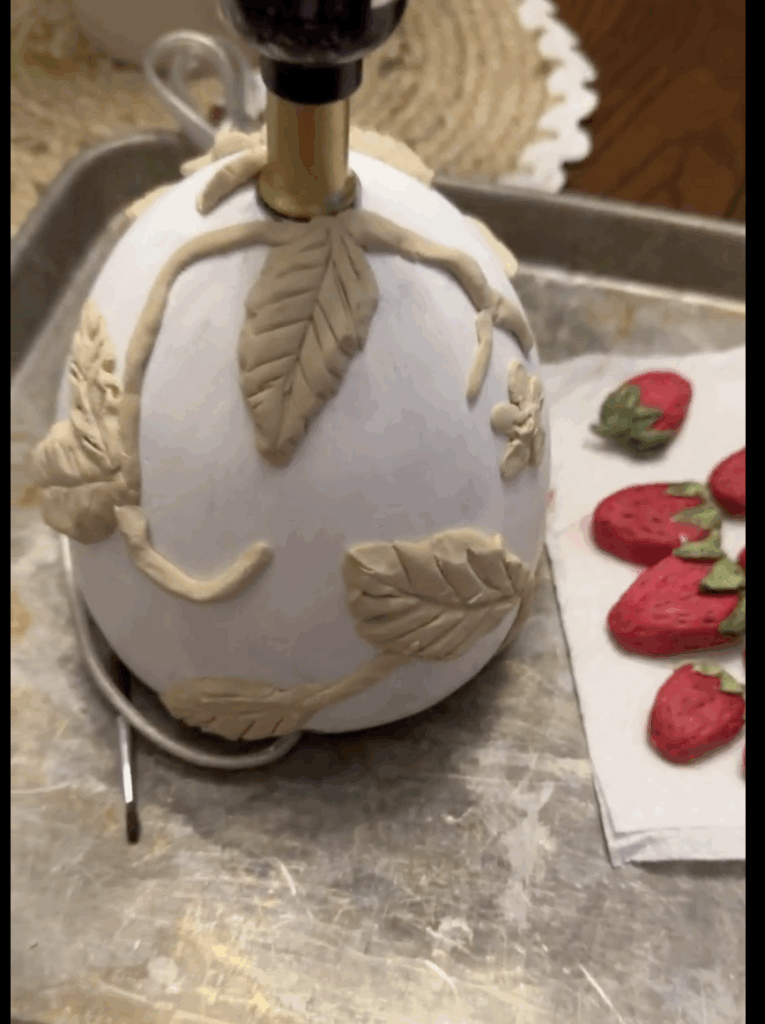 Lamp base covered in air dry clay vines and leaves with painted clay strawberries drying separately on a paper towel.