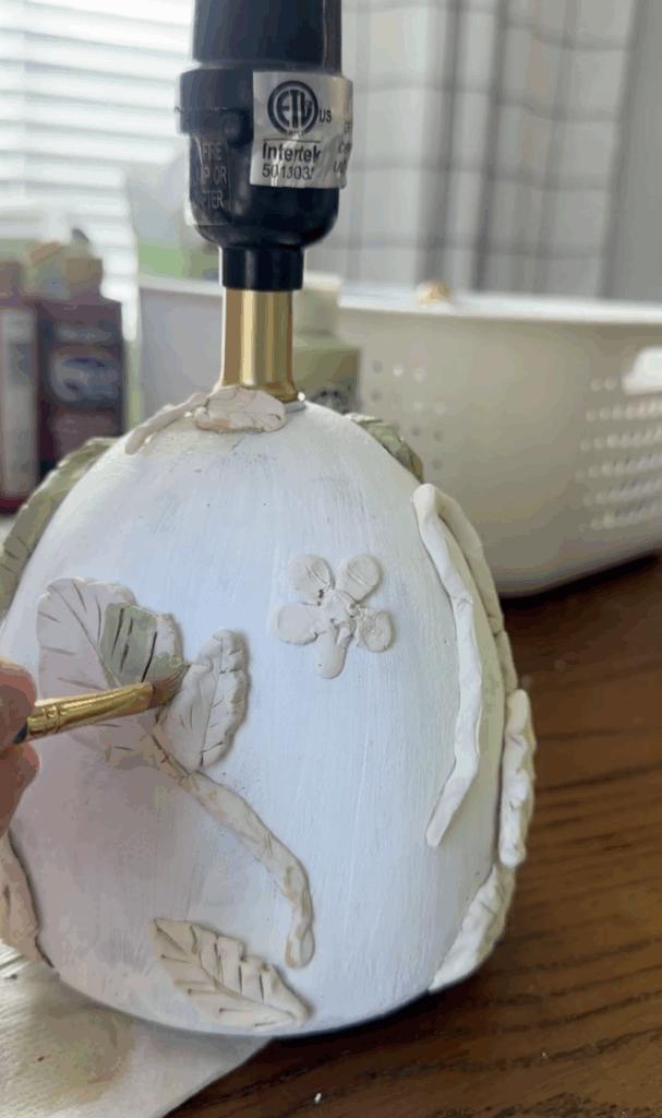 Painting green details onto sculpted clay leaves and vines attached to a white lamp base.