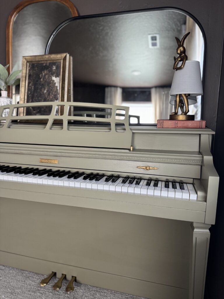 Finished painted piano in sage green with gold accents, styled with layered mirrors, vintage books, vase, and quirky gold rabbit lamp.
