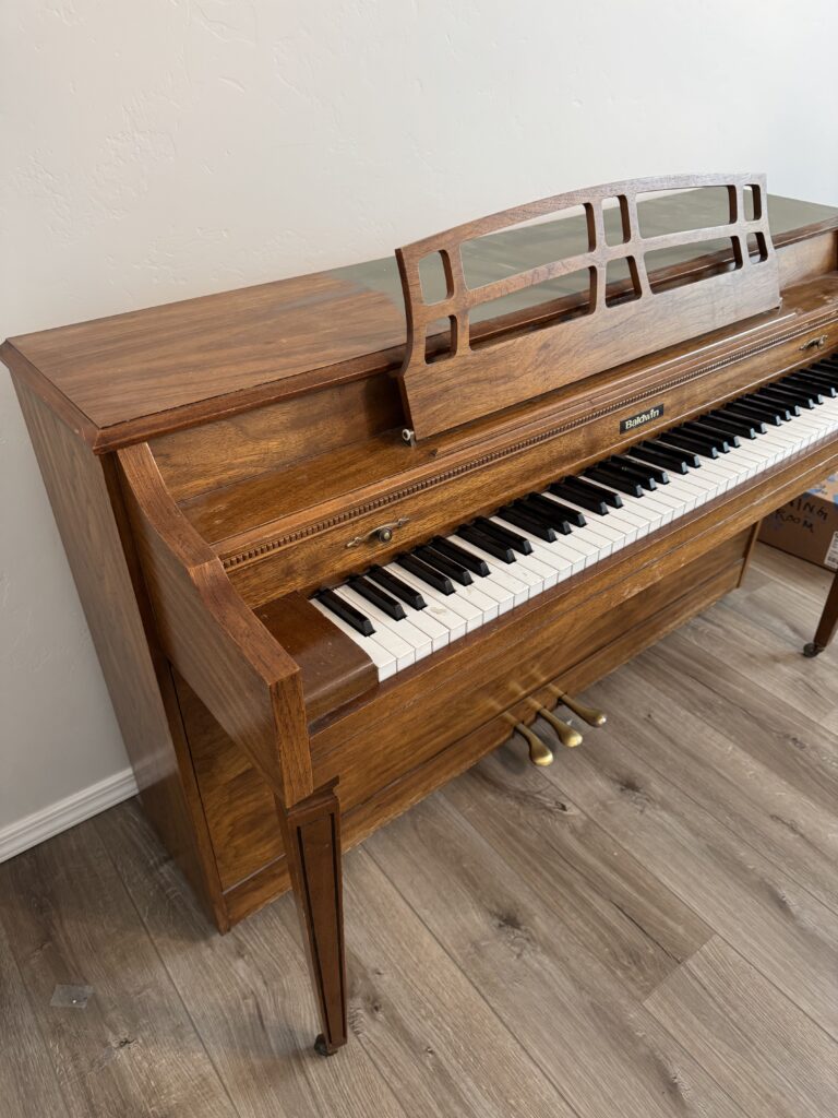 Side view of thrifted Baldwin piano before makeover, with wood grain and brass pedals visible.