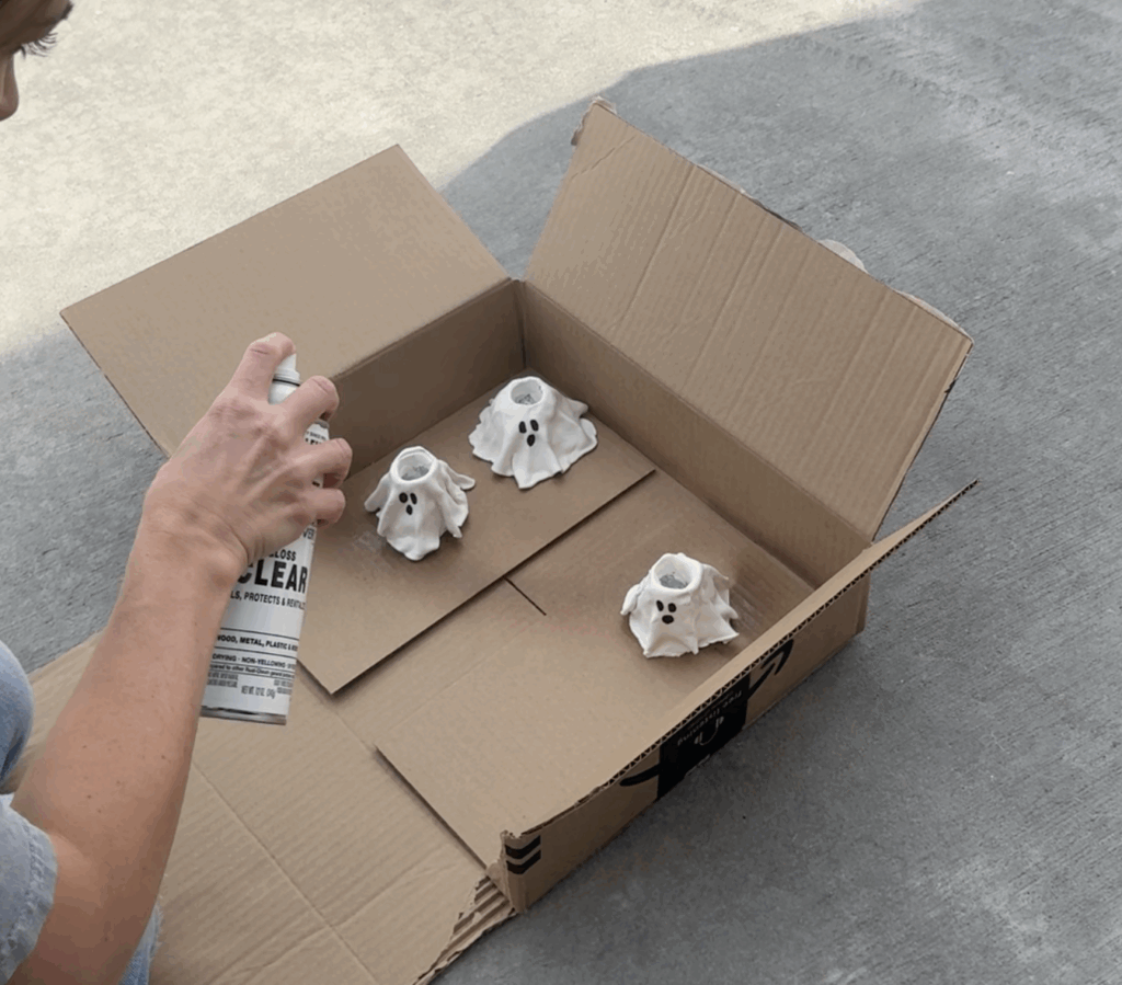 DIY ghost candlestick holders being sealed with clear spray paint in a cardboard box.